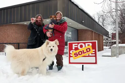 Two women standing outside a building in the snow. They have two dogs. 