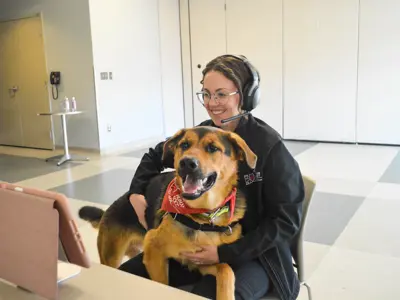 A woman with a headset has a dog on her lap while she teaches a class through a Zoom call. 